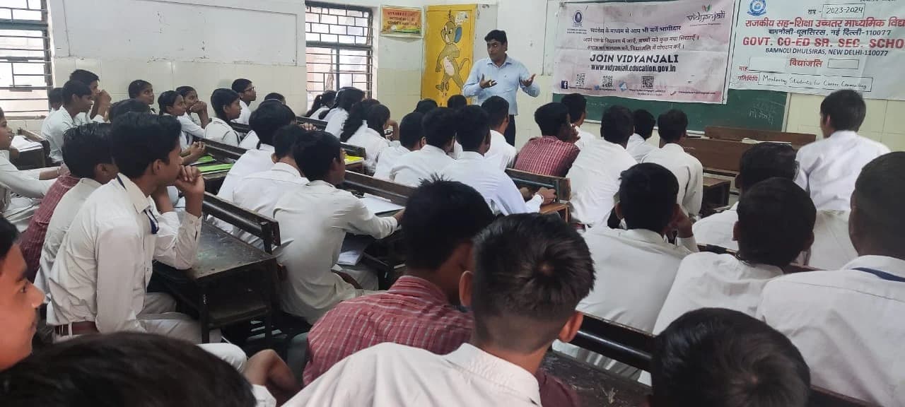 A mentor addressing a classroom of students under a 'Mentoring Students for Career Counselling' banner at a government school in Delhi.